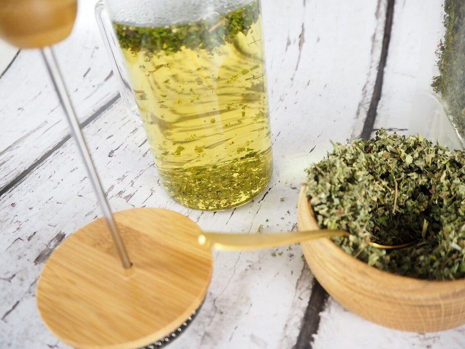 Infusion de feuilles de guimauve dans une tasse transparente avec une passoire à thé et une cuillère en bambou sur un fond en bois blanc.