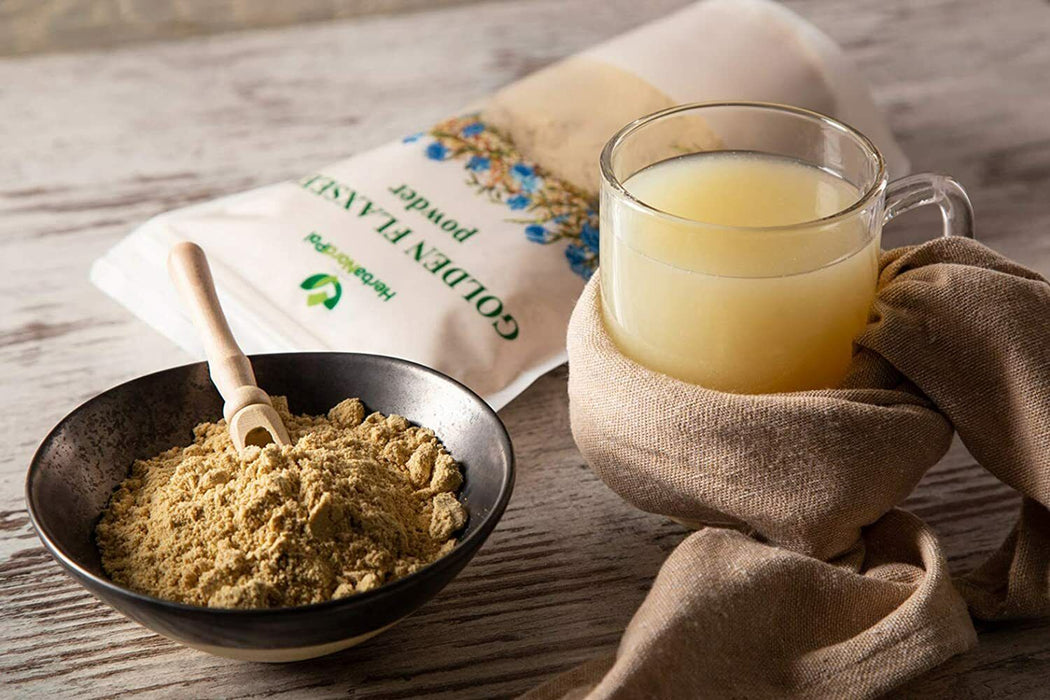 Poudre de graines de lin doré dans un bol sur une table en bois, à côté d'un verre de boisson jaune et d'un sachet de la marque HealthLink.