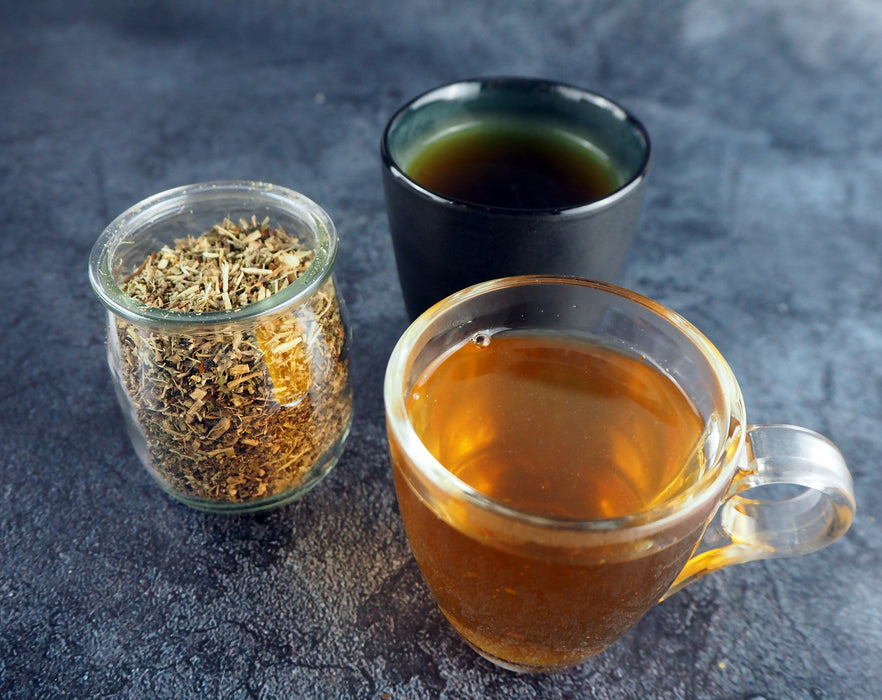 Deux tasses de thé aux herbes aux couleurs différentes, accompagnées de thé en vrac dans un bocal en verre sur un comptoir en ardoise.