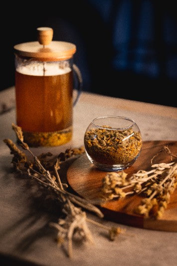 Théière moderne remplie de Herbal Tea Immuflos avec un verre de thé aux herbes à côté sur une table en bois.