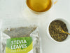 Thé de Stevia préparé avec des feuilles séchées, emballage transparent et cuillère de mesure sur fond blanc.