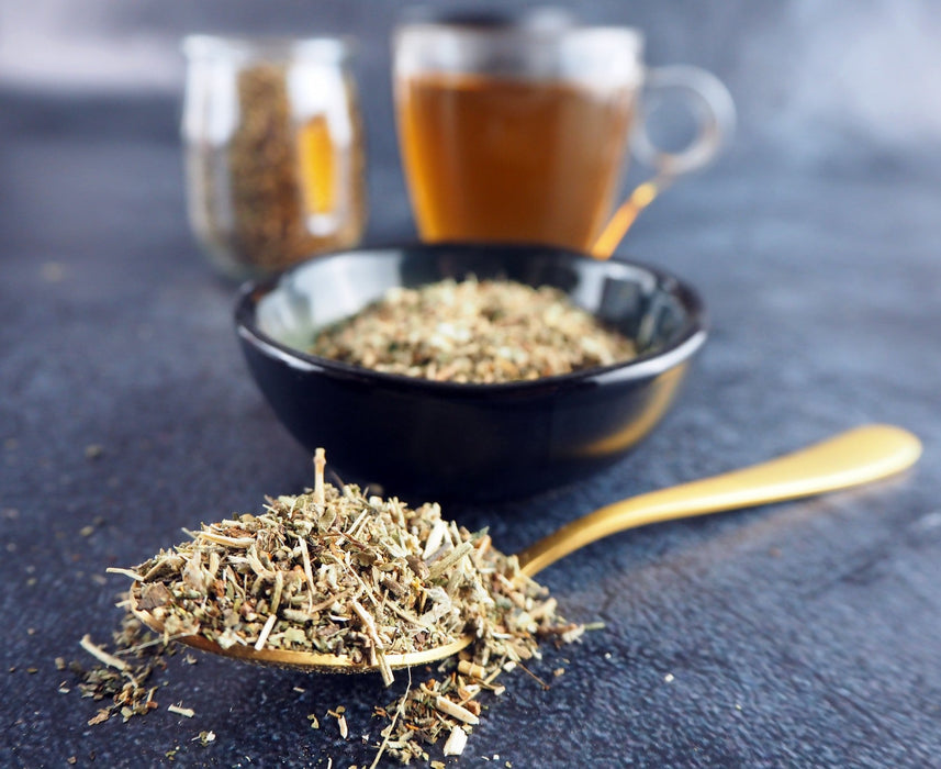 Cuillère dorée remplie de thé aux herbes sèches devant deux tasses de thé, prête pour la préparation d'une infusion relaxante.