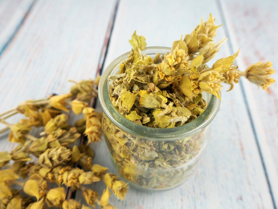 Bocal en verre rempli de Thé de montagne BIO Sideritis avec des fleurs séchées en arrière-plan, parfait pour une pause bien-être.