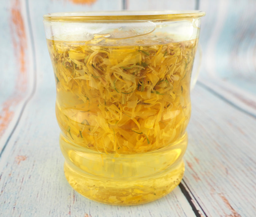 Une vue rapprochée d'une infusion dorée de souci dans une tasse transparente, évoquant un moment de calme et de relaxation avec ce thé aux herbes.L'emballage de Calendula officinalis dépeint une abondance de fleurs de souci, suggérant un thé aux multiples vertus pour les amateurs de tisanes naturelles.