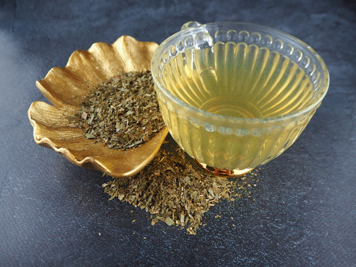 Un bol de feuilles de Ginkgo biloba à côté d'un thé infusé dans un bol en verre, mis en scène sur un fond texturé.