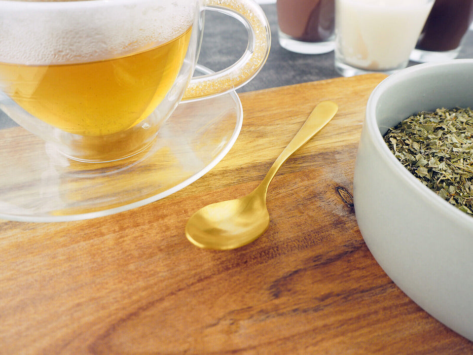 Infusion de thé de verveine citronnée dans une tasse transparente, accompagnée d'une cuillère dorée sur une table en bois.