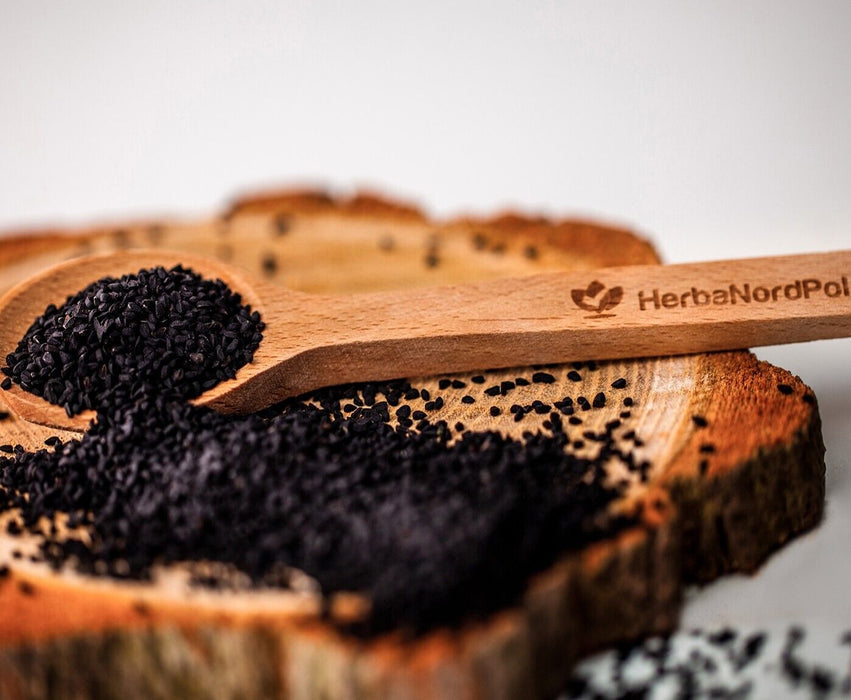 Cuillère en bois gravée HerbaNordPol reposant sur une planche de bois rustique, remplie de graines de Nigelle noire BIO fraîchement récoltées.